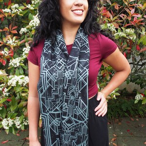 May include: A black scarf with a geometric design in shades of gray and teal. The scarf has a fringed edge and is draped around the neck. The person is wearing a maroon top and black pants.
