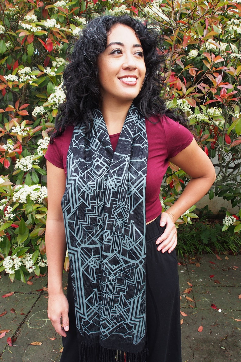 May include: A black scarf with a geometric design in shades of gray and teal. The scarf has a fringed edge and is draped around the neck. The person is wearing a maroon top and black pants.