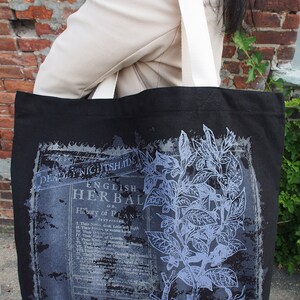 Op de afbeelding: Zwarte canvas tote bag met witte banden met een vintage botanische illustratie van Belladonna en tekst die "The English Herbal or History of Plants" luidt.