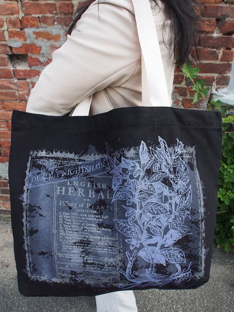Op de afbeelding: Zwarte canvas tote bag met witte banden met een vintage botanische illustratie van Belladonna en tekst die "The English Herbal or History of Plants" luidt.