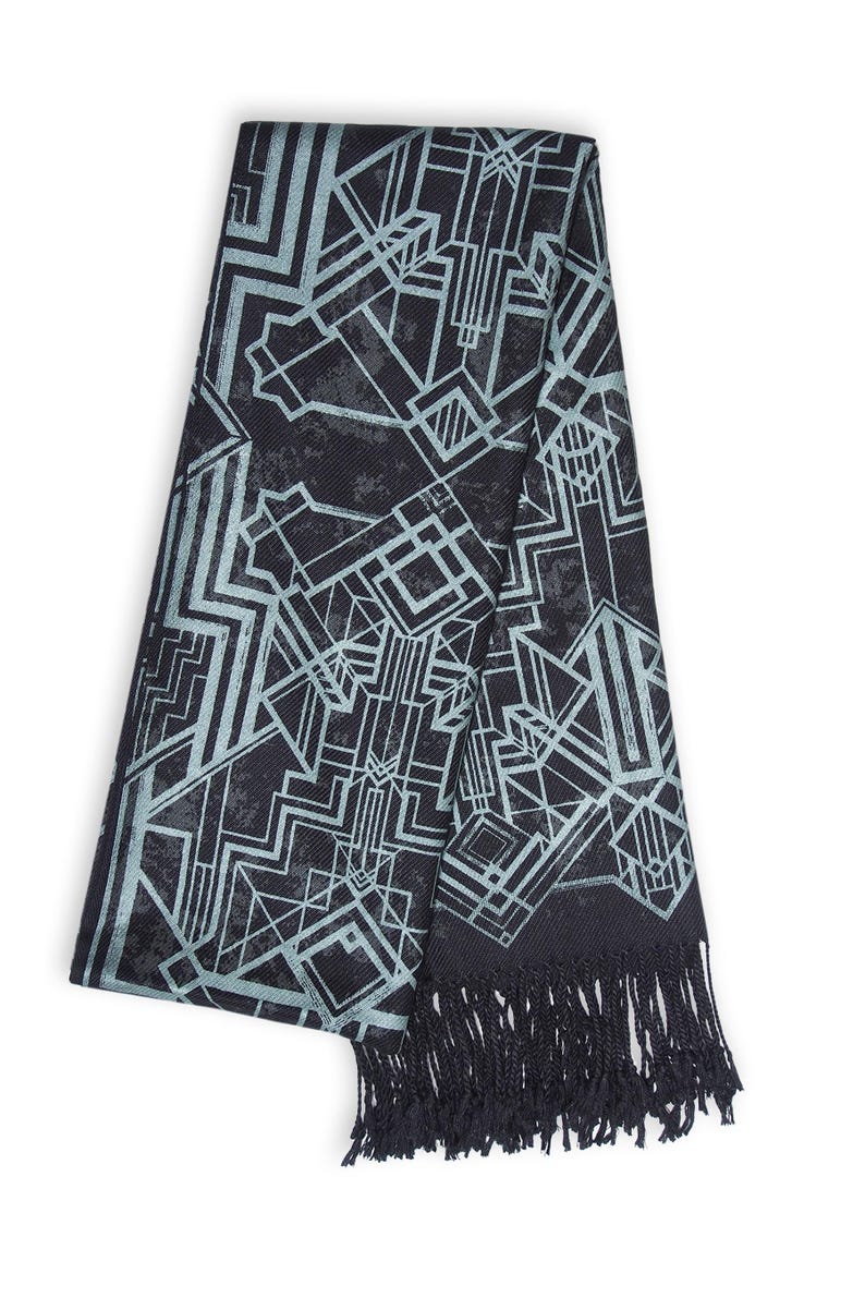 May include: A black scarf with a geometric Art Deco design in light blue. The scarf has a fringed edge. The pattern consists of lines and shapes, creating a modern and stylish accessory.