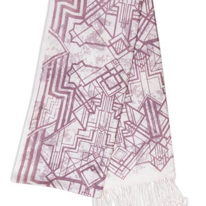 May include: A white and mauve geometric patterned scarf with a fringed edge. The design features an Art Deco style with angular shapes and lines. The scarf is folded, showcasing the pattern and texture.