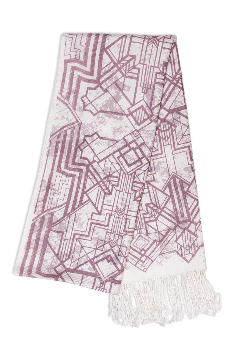 May include: A white and mauve geometric patterned scarf with a fringed edge. The design features an Art Deco style with angular shapes and lines. The scarf is folded, showcasing the pattern and texture.