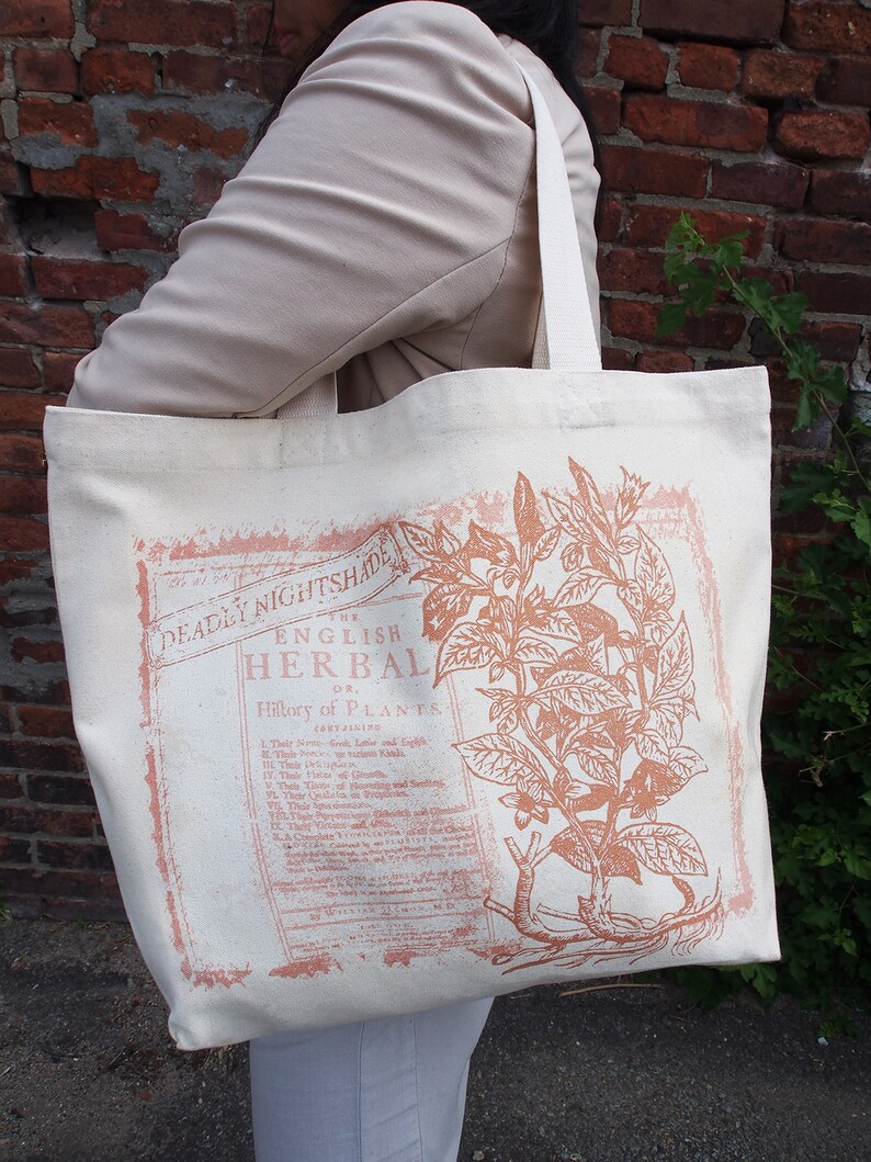 Op de afbeelding: Een witte canvas tote bag met een vintage botanische illustratie van een plant. De illustratie is in roze en bruine tinten. De tekst op de tas luidt "The English Herbal or History of Plants".