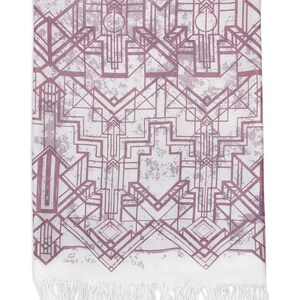 May include: A rectangular white scarf with a geometric Art Deco design in shades of mauve and gray. The scarf has a distressed, textured appearance and is finished with a white fringe along the bottom edge.