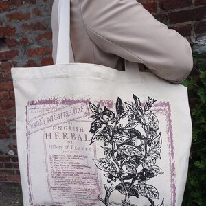Op de afbeelding: Een witte canvas tote bag met zwarte banden. De tas heeft een vintage botanische illustratie van een plant met de tekst "The English Herbal or History of Plants" gedrukt op de voorkant.