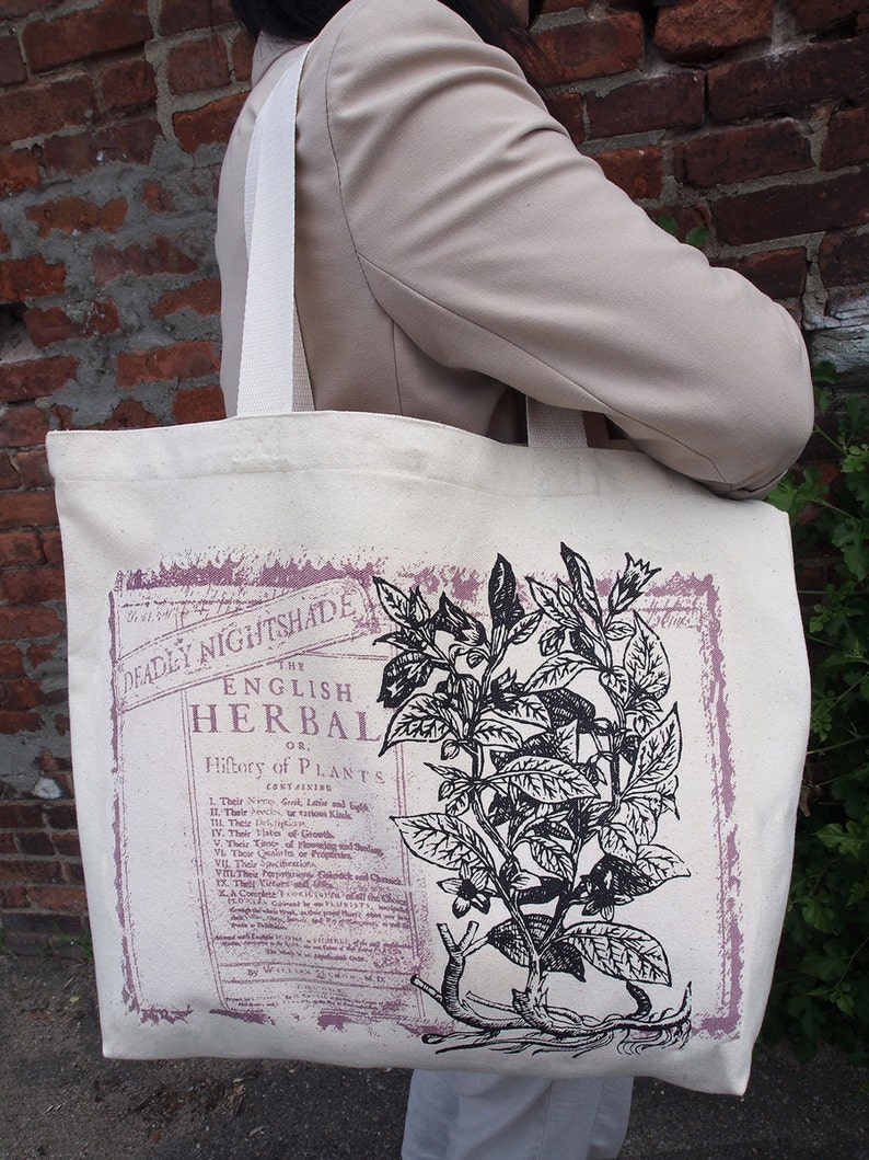 Op de afbeelding: Een witte canvas tote bag met zwarte banden. De tas heeft een vintage botanische illustratie van een plant met de tekst "The English Herbal or History of Plants" gedrukt op de voorkant.