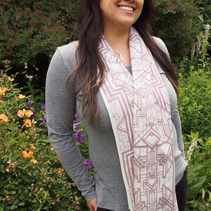 May include: A light-colored scarf with a geometric design in shades of mauve and white. The scarf has a fringed edge and is draped over a gray long-sleeved shirt. The background features lush green foliage and colorful flowers.