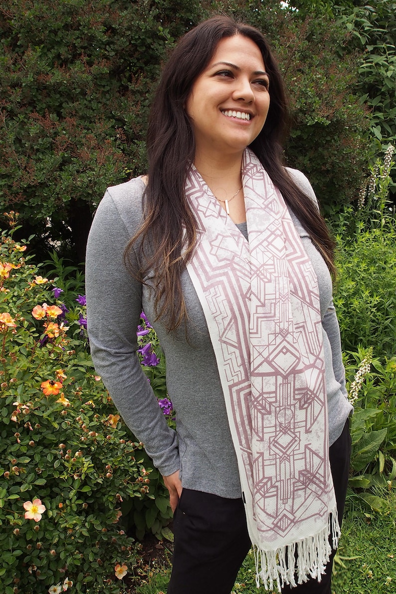 May include: A light-colored scarf with a geometric design in shades of mauve and white. The scarf has a fringed edge and is draped over a gray long-sleeved shirt. The background features lush green foliage and colorful flowers.