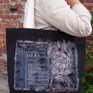 Op de afbeelding: Zwarte tote bag met een witte band en een vintage botanische illustratie van een plant met de tekst "The English Herbal or History of Plants" erop gedrukt.