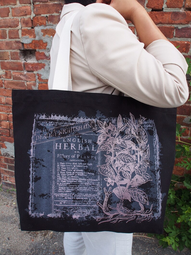 Op de afbeelding: Zwarte tote bag met een witte band en een vintage botanische illustratie van een plant met de tekst "The English Herbal or History of Plants" erop gedrukt.