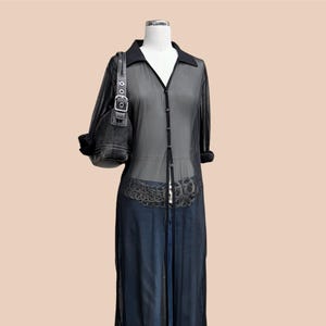 May include: A black sheer button-down duster shirt with a collar, paired with a dark denim skirt. A black handbag with a silver buckle hangs on the mannequin's arm. The duster shirt is long and reaches the ankles.