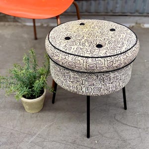 May include: A round ottoman featuring a cream-colored fabric with a black abstract geometric pattern. The top is tufted with four black buttons and finished with black piping. It stands on four thin, straight black metal legs.