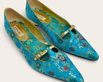 May include: A pair of pointed-toe flats in bright turquoise fabric with a multi-colored floral pattern. Each shoe features a thin gold strap across the instep with a small gold buckle. The interior is lined in gold, with a label reading ISAAC MADE IN ITALY.