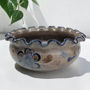 May include: A round ceramic bowl with a ruffled, scalloped rim featuring a dark blue edge. The exterior has a speckled beige and brown glaze decorated with hand-painted dark blue and brown floral motifs and small textured hatch patterns.