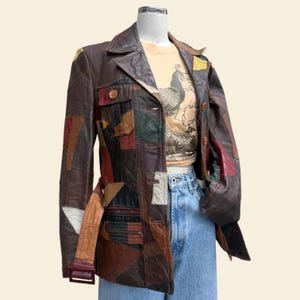 May include: A patchwork leather jacket featuring a multi-colored design with shades of brown, tan, red, green, and black. The jacket includes a collared neckline, a button-front closure, a belted waist, and two chest pockets with button flaps.
