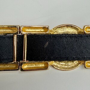 Puede incluir: Un cintur&oacute;n negro con eslabones met&aacute;licos dorados rectangulares y circulares. El cintur&oacute;n consiste en una correa negra lisa que pasa a trav&eacute;s de una serie de componentes geom&eacute;tricos de color dorado, incluyendo marcos rectangulares y una pieza central.