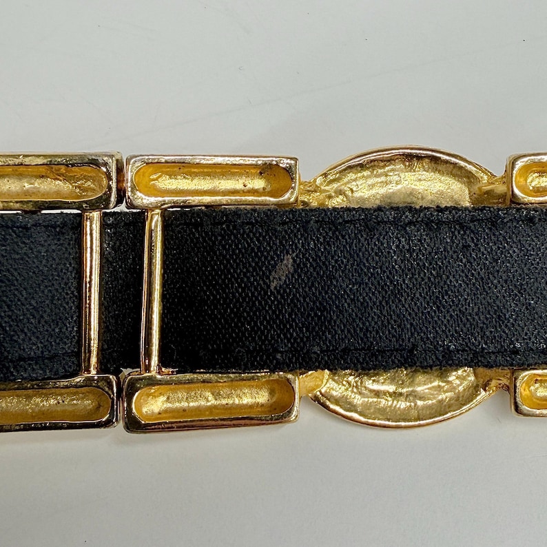 Puede incluir: Un cintur&oacute;n negro con eslabones met&aacute;licos dorados rectangulares y circulares. El cintur&oacute;n consiste en una correa negra lisa que pasa a trav&eacute;s de una serie de componentes geom&eacute;tricos de color dorado, incluyendo marcos rectangulares y una pieza central.