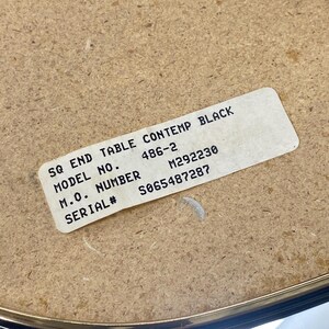 May include: A close-up of a label on a square end table. The label reads "SQ END TABLE CONTEMP BLACK" with the model number "486-2", M.O. number "M292230", and serial number "S065487287". The table has a gold-colored trim.