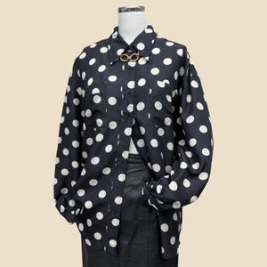 May include: A long-sleeved button-down shirt in black with a white polka dot pattern. The shirt features a classic collar, a front button placket, two chest pockets, and a gold-toned metal link detail at the neckline. It is displayed on a mannequin.