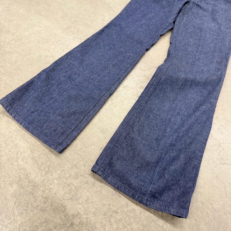 May include: A pair of blue denim bell-bottom trousers. The trousers have a high waist and a wide flare from the knee down. The denim is a medium wash, and the trousers appear to be vintage.