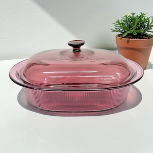 May include: A vintage, ruby-colored glass casserole dish with a matching lid. The dish has a ribbed design and a rounded shape. The lid features a small, round handle. A small potted plant is in the background.