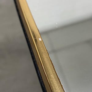 May include: Close-up of a gold-colored, rectangular frame with a dark blue edge. The frame has a reflective surface and appears to be slightly angled. The background is a blurred, neutral color.