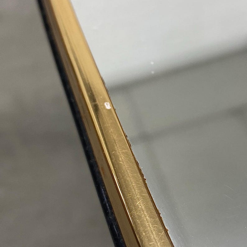 May include: Close-up of a gold-colored, rectangular frame with a dark blue edge. The frame has a reflective surface and appears to be slightly angled. The background is a blurred, neutral color.