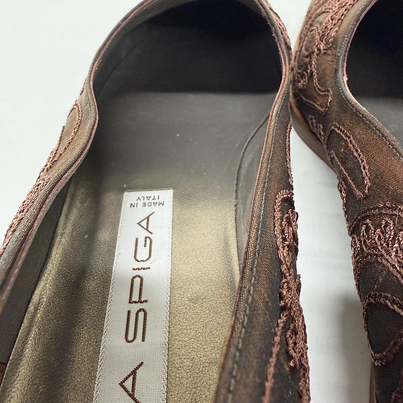 May include: Close-up of a pair of brown flats with detailed embroidery. The shoes have a pointed toe and a low heel. The interior is light brown, with the brand name "A SPIGA" visible. Made in Italy.