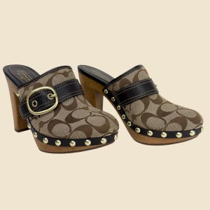 May include: A pair of Coach clogs with a brown and tan monogram pattern. The clogs have wooden heels and soles, black leather straps, and gold-colored studs around the sole. A gold buckle secures the strap.