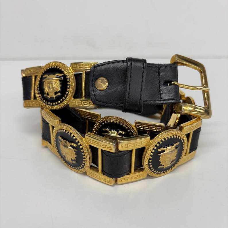 Puede incluir: Un cintur&oacute;n de cuero negro con herrajes de metal en tono dorado. El cintur&oacute;n est&aacute; adornado con medallones circulares que muestran un perfil de cabeza en relieve dorado sobre fondo negro, unidos por eslabones rectangulares con grecas.