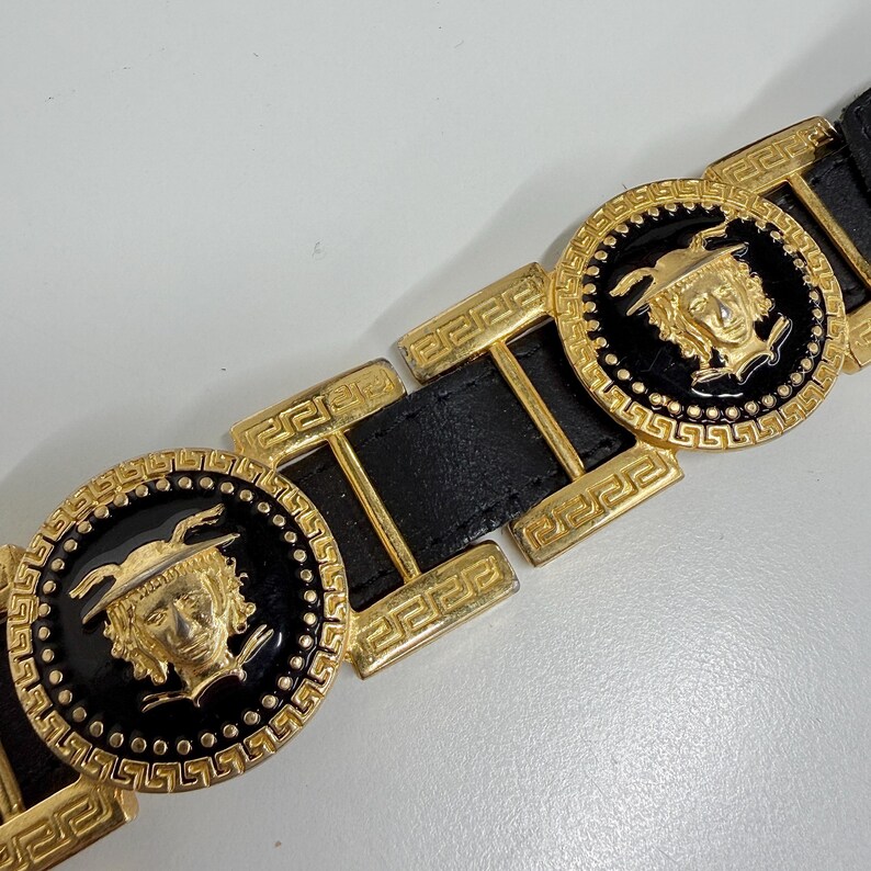 Puede incluir: Un cintur&oacute;n de cuero negro con eslabones met&aacute;licos dorados repetidos. El dise&ntilde;o incluye medallones circulares con un motivo de rostro dorado en relieve sobre fondo negro, rodeados por un borde con patr&oacute;n de greca en el metal.