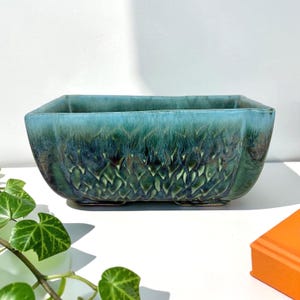 May include: A rectangular ceramic planter featuring a reactive glaze in shades of teal, deep blue, and forest green. The exterior surface displays a textured, vertical drip pattern that transitions from a lighter turquoise rim to a dark base.