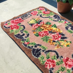 May include: A rectangular rug with a floral design. The rug features a light pink center with a brown border. The floral pattern includes red, pink, yellow, and green flowers with dark blue accents. The rug is placed on a concrete surface.