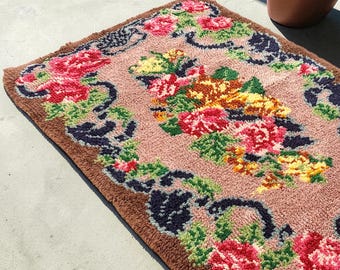 May include: A rectangular rug with a floral design. The rug features a light pink center with a brown border. The floral pattern includes red, pink, yellow, and green flowers with dark blue accents. The rug is placed on a concrete surface.