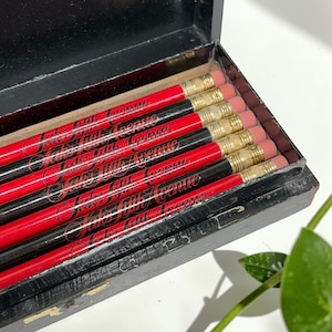 May include: A black wooden box filled with pencils. The pencils are red and black with gold-colored metal bands and pink erasers. The text "Saks Fifth Avenue" is printed on the pencils in a cursive font.