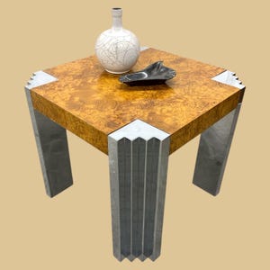 May include: A square side table with a burl wood top and geometric metal legs. The table features a decorative white vase with black line art and a black dish. The table's dimensions are approximately 24 inches square.