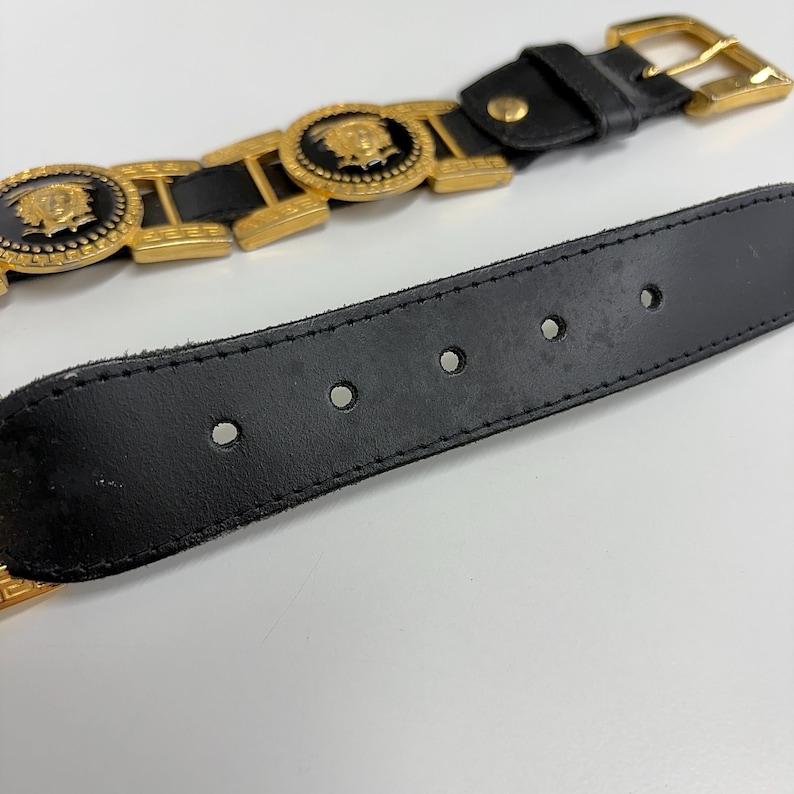 Puede incluir: Un cintur&oacute;n de cuero negro con herrajes de metal en tono dorado. El cintur&oacute;n incluye medallones circulares con un emblema central de cabeza de Medusa dorada sobre fondo negro, unidos por eslabones dorados. Tiene una hebilla dorada y cinco agujeros.