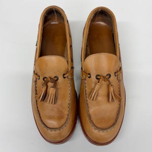 May include: A pair of tan leather loafers featuring a moccasin-style toe, dark brown metal eyelets, and decorative leather tassels tied at the front. The shoes have a smooth finish, visible stitching along the vamp, and a dark brown sole edge.