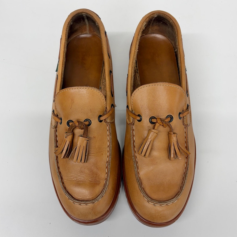 May include: A pair of tan leather loafers featuring a moccasin-style toe, dark brown metal eyelets, and decorative leather tassels tied at the front. The shoes have a smooth finish, visible stitching along the vamp, and a dark brown sole edge.