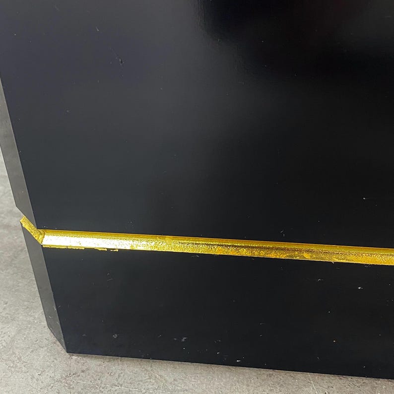May include: A black, geometric box with a gold-colored trim. The box has a glossy finish and a modern design. The gold trim accents the edges of the box, creating a striking contrast.