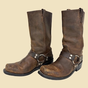May include: A pair of brown leather motorcycle boots. The boots have a harness strap with silver-tone hardware and a square toe. The leather shows wear and tear, with scuff marks on the toes and sides. The soles are black.
