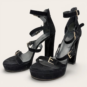 May include: A pair of black suede platform sandals featuring high block heels, multiple adjustable straps across the foot and ankle, and gold-toned metal buckles. The design includes a rounded toe, a sturdy heel base, and a secure ankle strap.