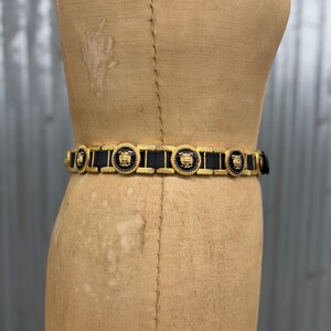 Puede incluir: Un cintur&oacute;n de cuero negro con eslabones de metal dorado. El cintur&oacute;n est&aacute; adornado con medallones circulares dorados, cada uno con un motivo de rostro dorado en relieve sobre un fondo negro, alternando con conectores rectangulares de metal dorado.