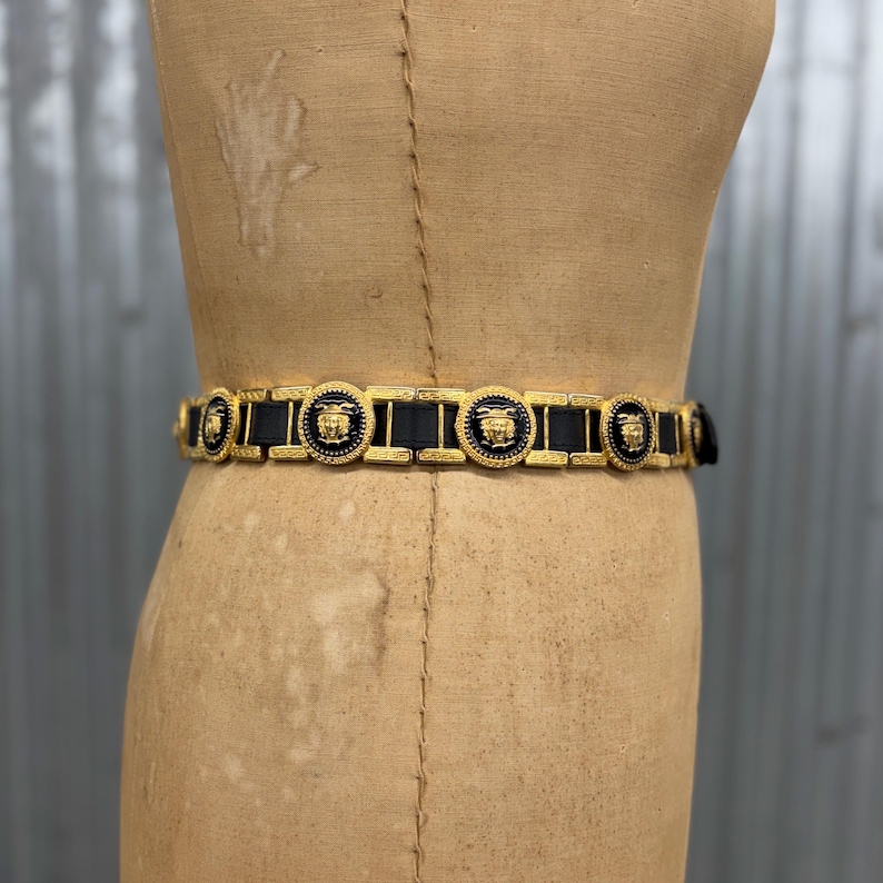 Puede incluir: Un cintur&oacute;n de cuero negro con eslabones de metal dorado. El cintur&oacute;n est&aacute; adornado con medallones circulares dorados, cada uno con un motivo de rostro dorado en relieve sobre un fondo negro, alternando con conectores rectangulares de metal dorado.