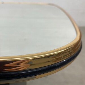 May include: Close-up of a table corner with a gold-colored metal frame and a black trim. The table has a clear glass top. The design is a classic, elegant style.
