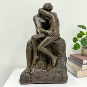 May include: A bronze sculpture depicting two figures embracing. The figures are nude and seated on a textured base. The sculpture has a dark brown patina and is displayed on a white surface.