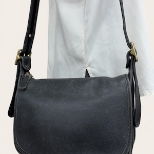 May include: A black leather shoulder bag featuring a rounded flap closure, a long adjustable strap, and antique brass-toned metal hardware. The bag has a smooth texture with visible natural grain and a zipper pull tab on the side.
