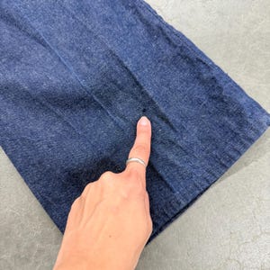 May include: Close-up of dark blue denim fabric. A finger with a silver ring points to a small dark spot on the denim. The fabric is folded, showing the texture and colour variations of the material.