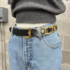 Puede incluir: Un cintur&oacute;n de cuero negro con una hebilla dorada y eslabones met&aacute;licos dorados decorativos con medallones negros circulares que muestran un emblema de cabeza de Medusa dorada en relieve, usado sobre jeans azul claro y un su&eacute;ter de punto gris.